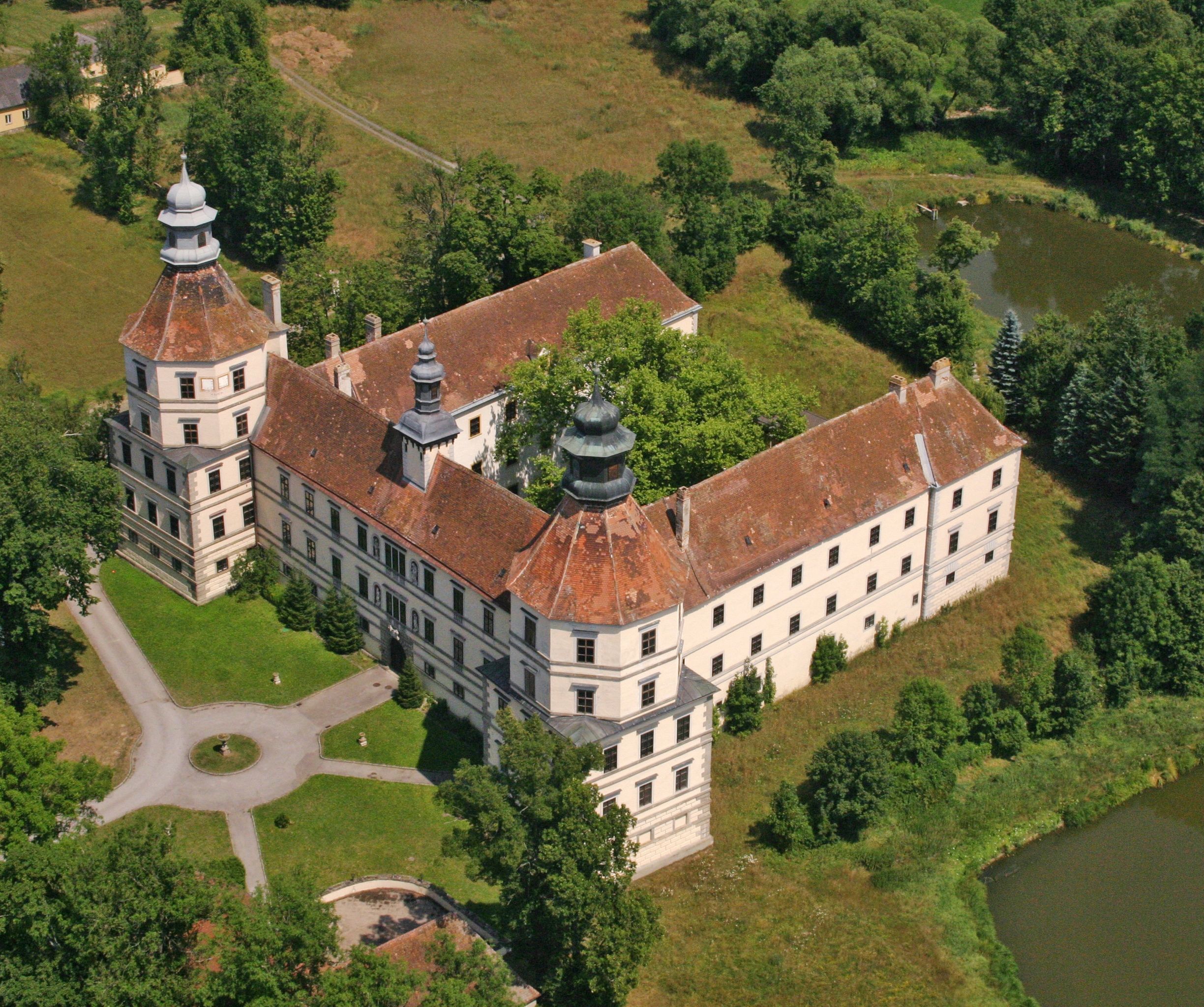 Letecký pohľad na historický hrad s dvoma vežami a vnútorným nádvorím, obklopený stromami a rybníkom.
