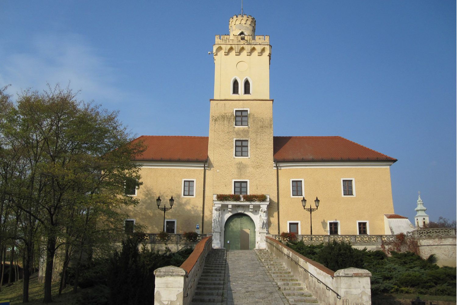 Hrad so žltou fasádou a červenou strechou, centrálna veža, široké kamenné schodisko, modrá obloha.