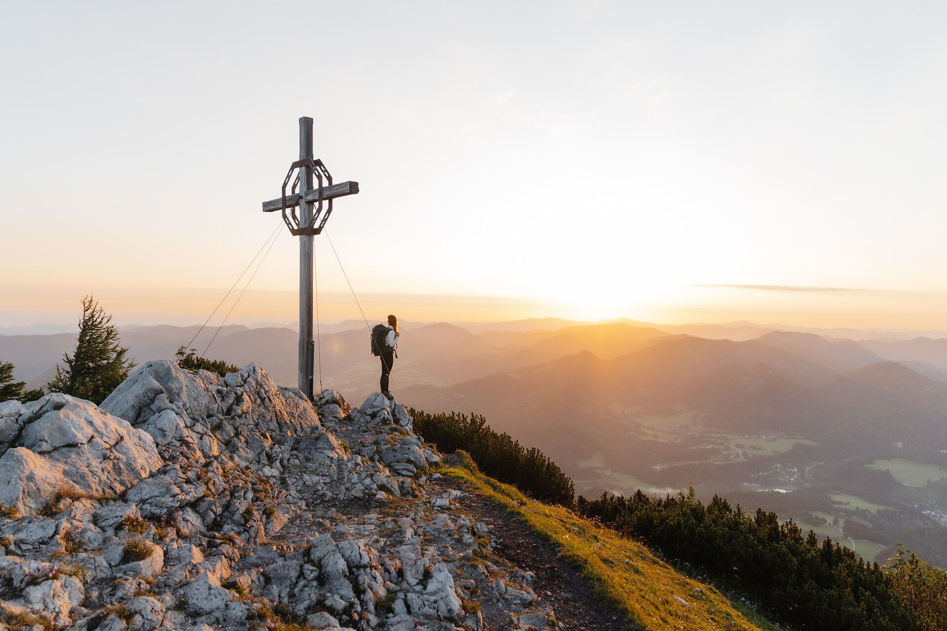 Die goldenen Strahlen der Abendsonne tauchen die majestätischen Berge in ein warmes Licht, während ein Wanderer auf dem Gipfel steht und die atemberaubende Aussicht genießt. Umgeben von sanften Hügeln und dichten Wäldern, vermittelt dieser Ort ein Gefühl von Freiheit und Abenteuer.