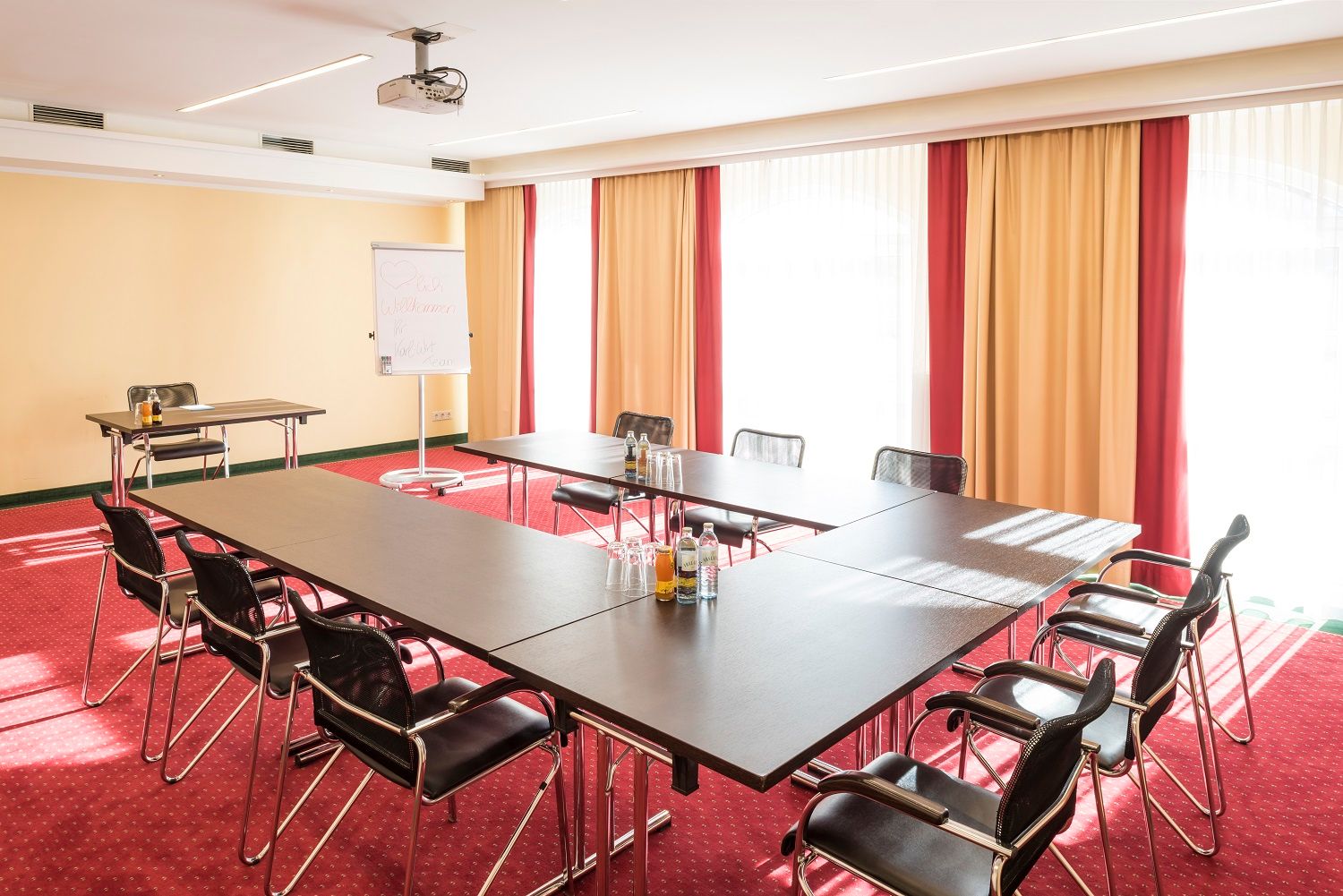 Konferenčná miestnosť so stolom v tvare U, stoličkami, flipchartom a projektorom.