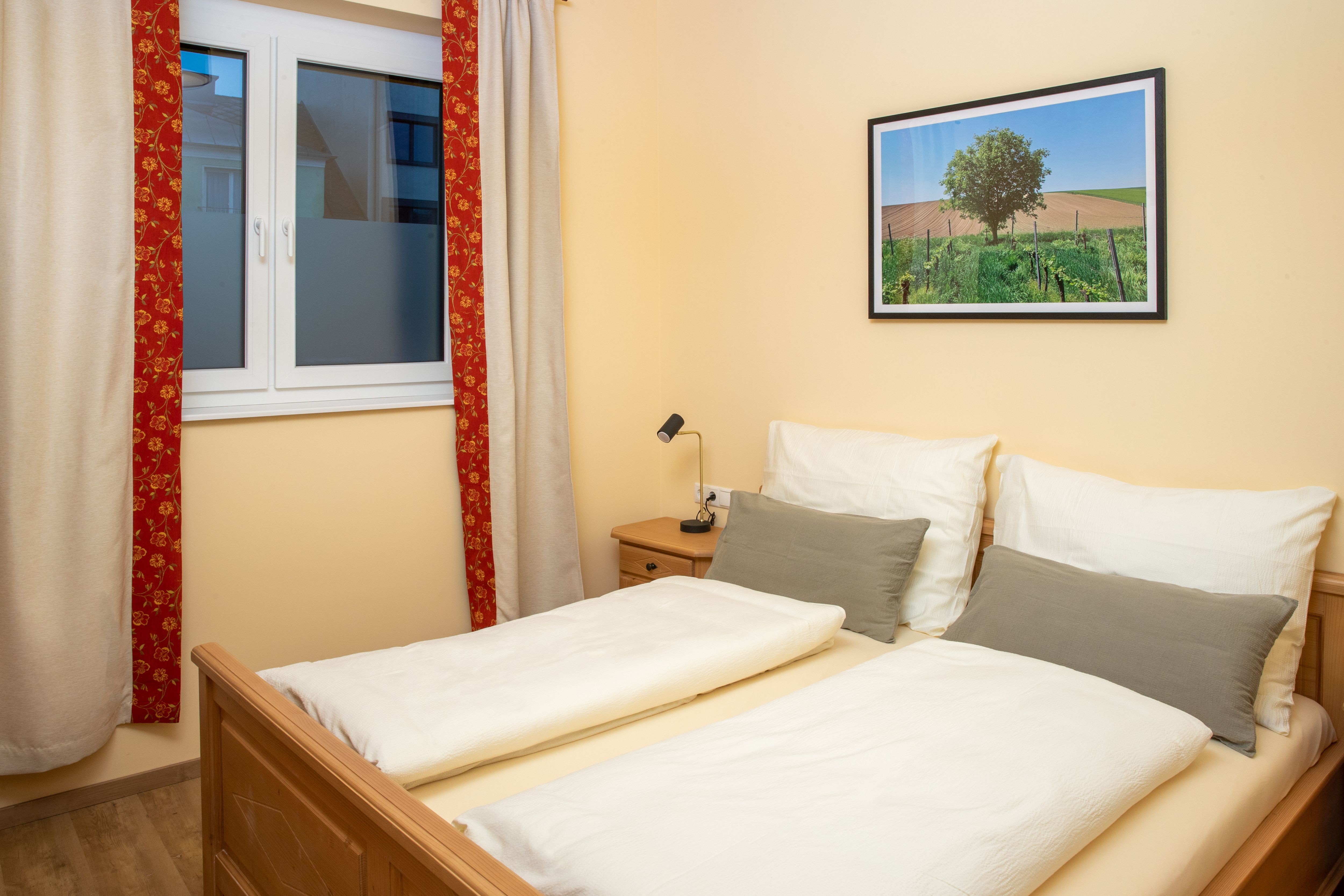 Útulná dvojlôžková izba s manželskou posteľou, oknom so závesmi a maľbou krajiny na stene.