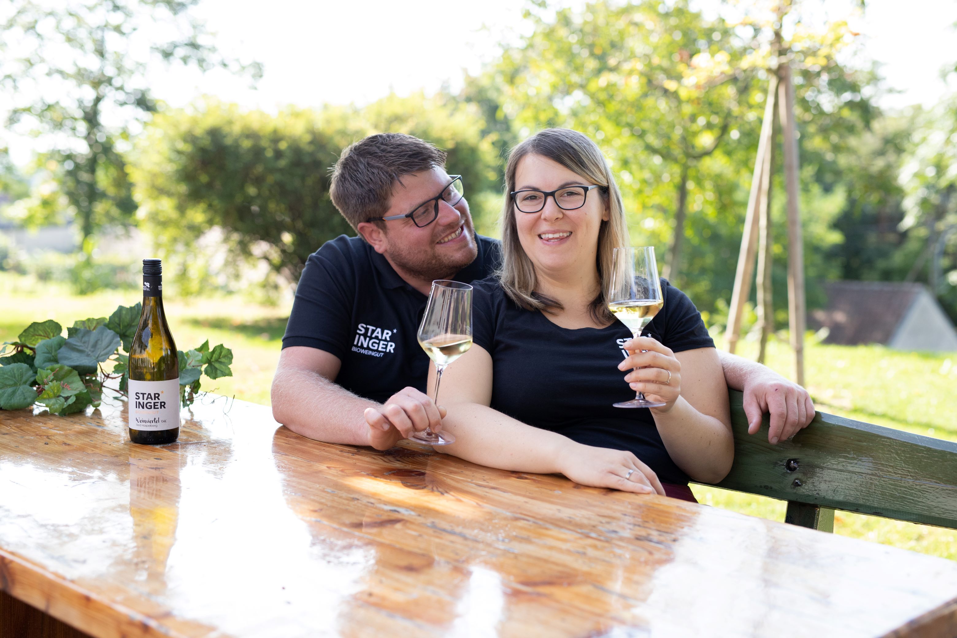 Pri drevenom stole vonku sedí dvojica s pohármi vína. Na stole je fľaša vína s nápisom "Staringer".