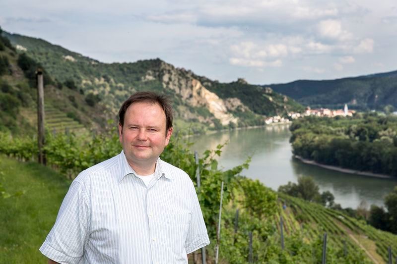 Muž stojaci vo vinici s riekou a kopcami v pozadí.