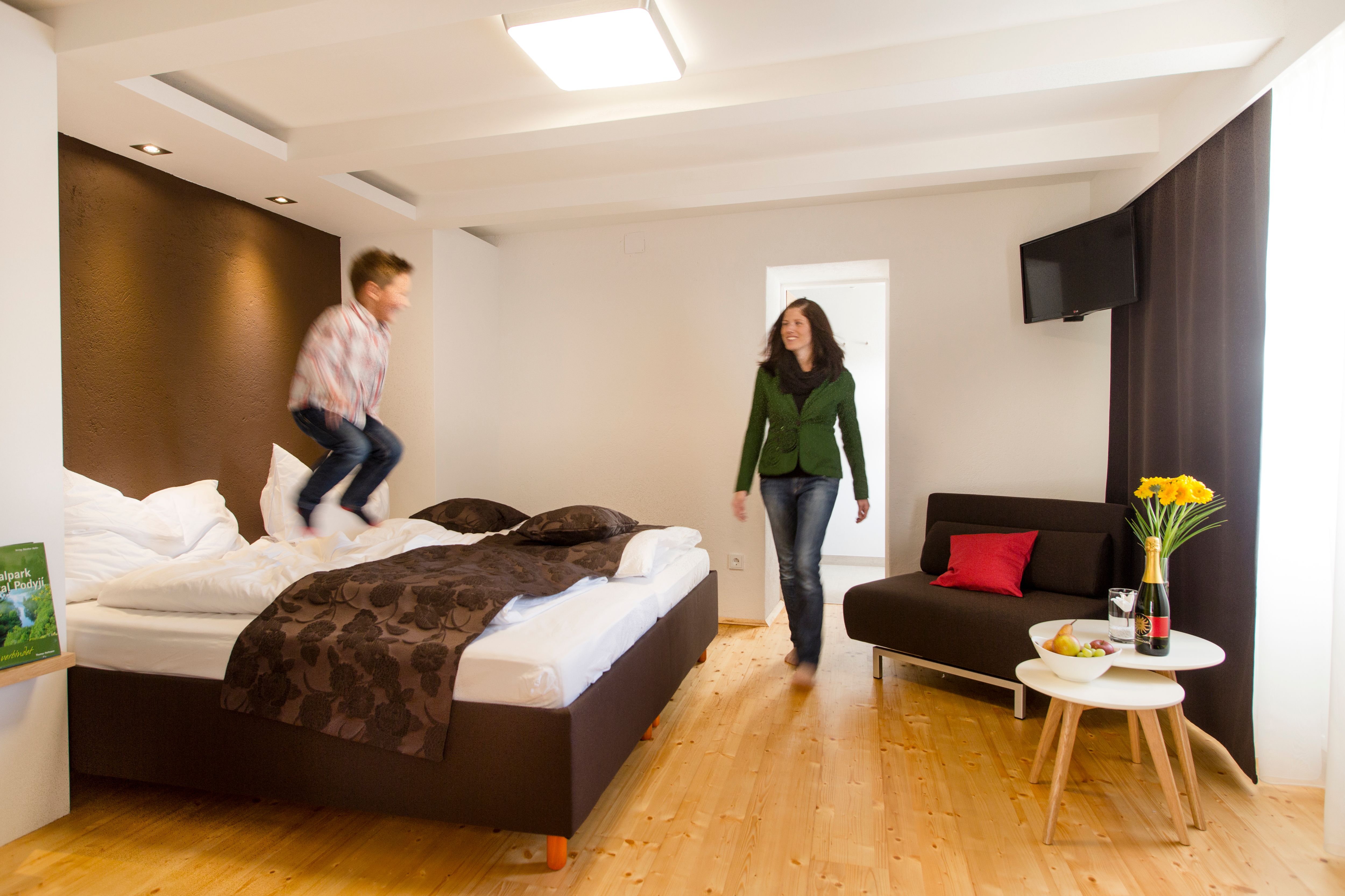 Dieťa skáče na posteli v modernej hotelovej izbe, zatiaľ čo žena sa usmieva. Vedľa stojí stôl s kvetmi a vínom.