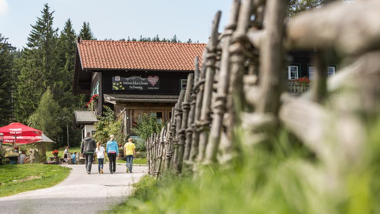 Traja ľudia kráčajú k horskému hostincu, ktorý je obklopený dreveným plotom a stromami.