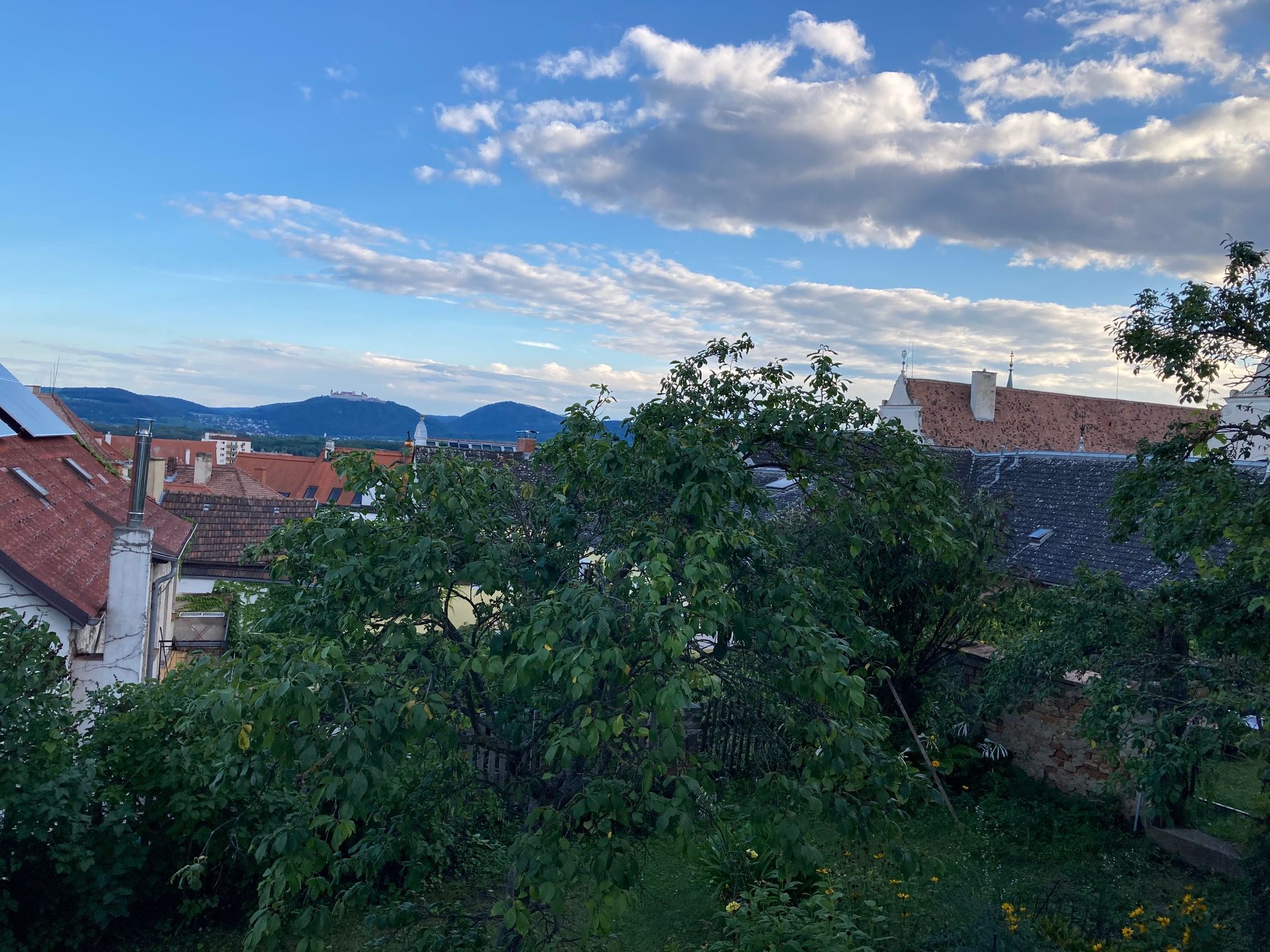 Pohľad cez strechy a stromy na kopce a modrú oblohu s mrakmi.