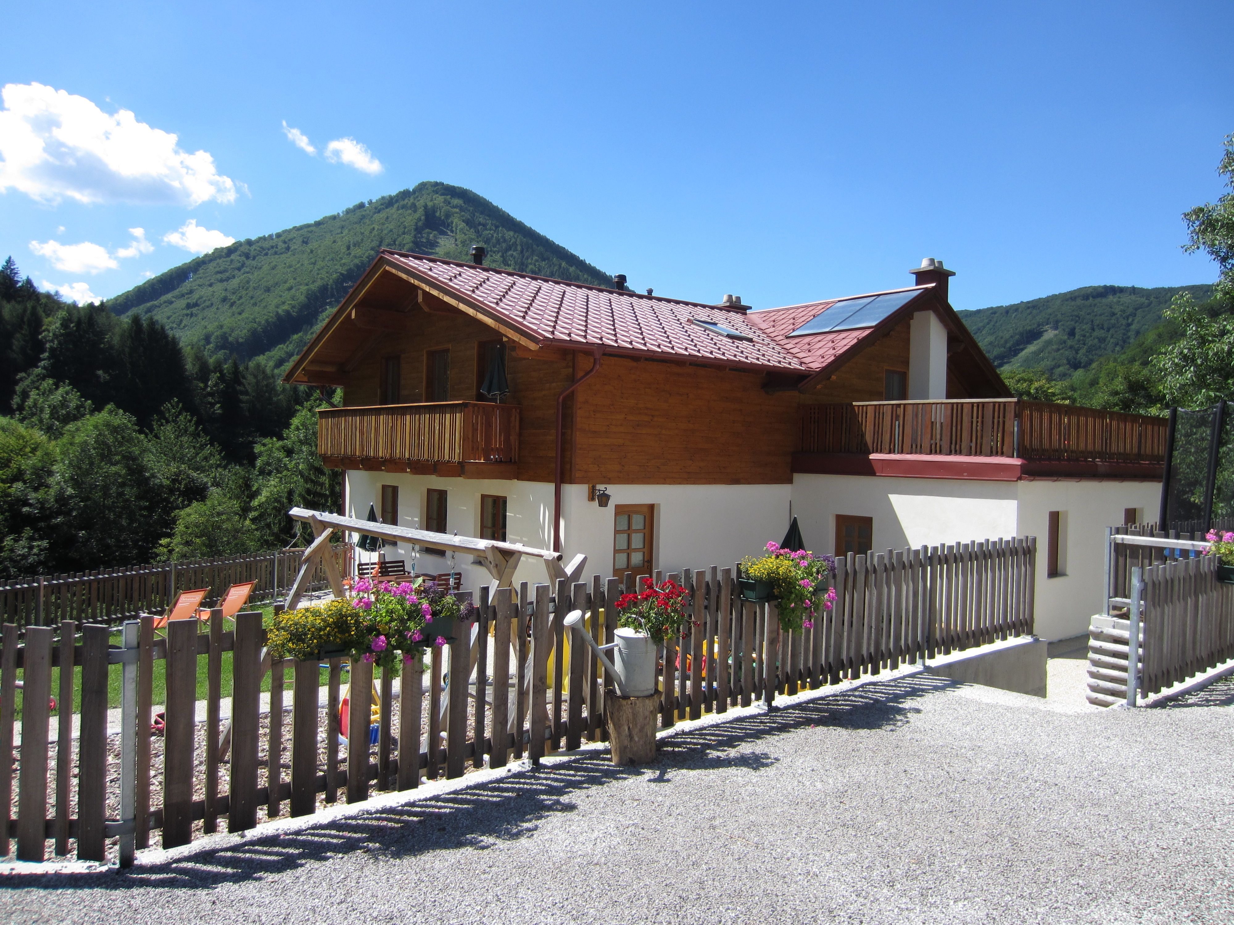 Dvojpodlažný dom s dreveným balkónom a záhradou, obklopený horami a stromami.