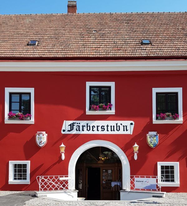Červená budova s nápisom "Färberstub'n" a rozkvitnutými kvetmi v oknách.