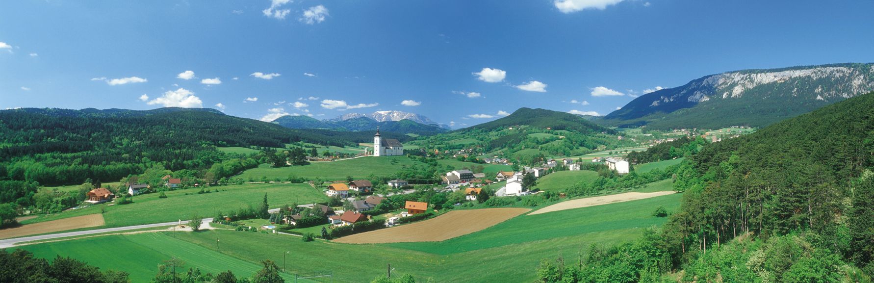 Panoramatický pohľad na Höflein s kostolom, zelenými poliami a horami v pozadí.