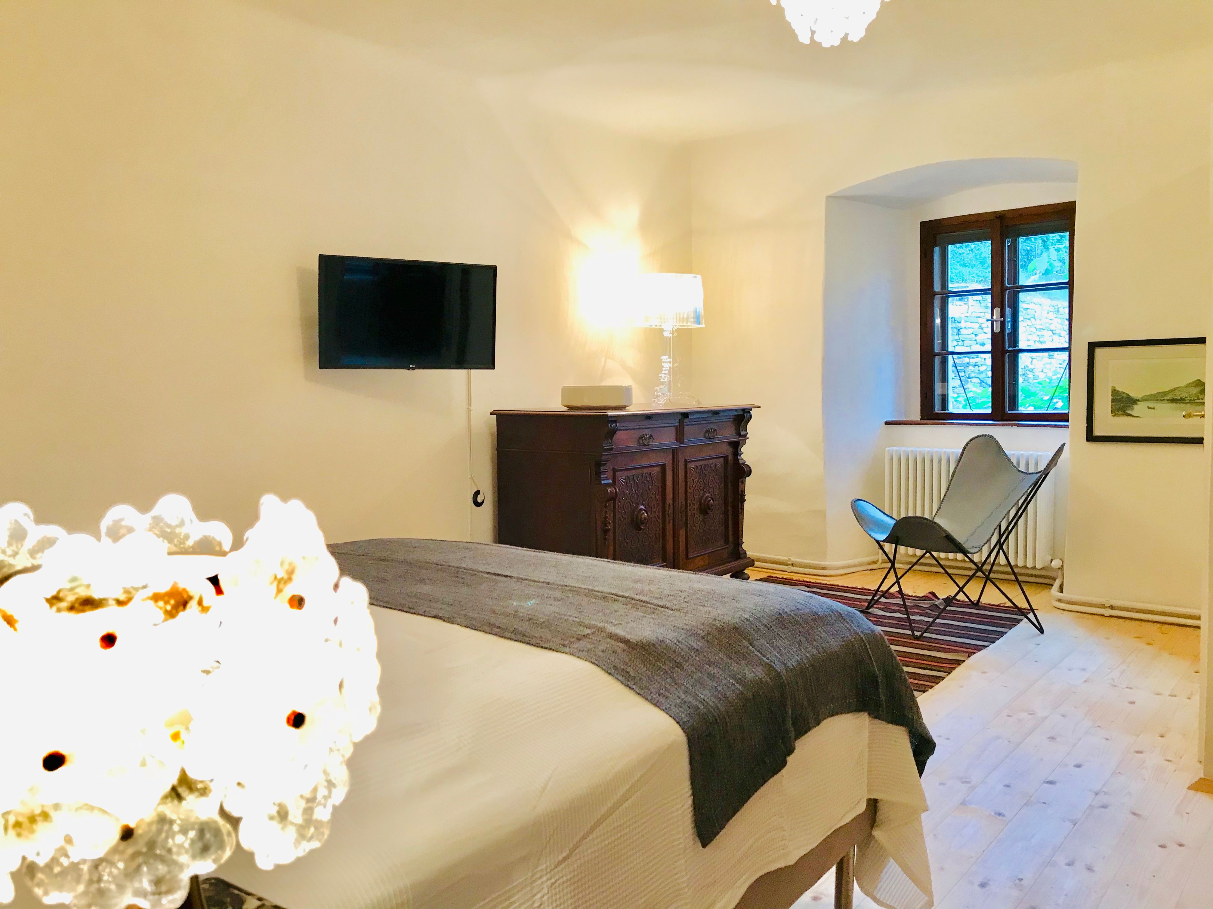 Útulná spálňa s posteľou, starožitnou komodou, kreslom a nástenným TV.