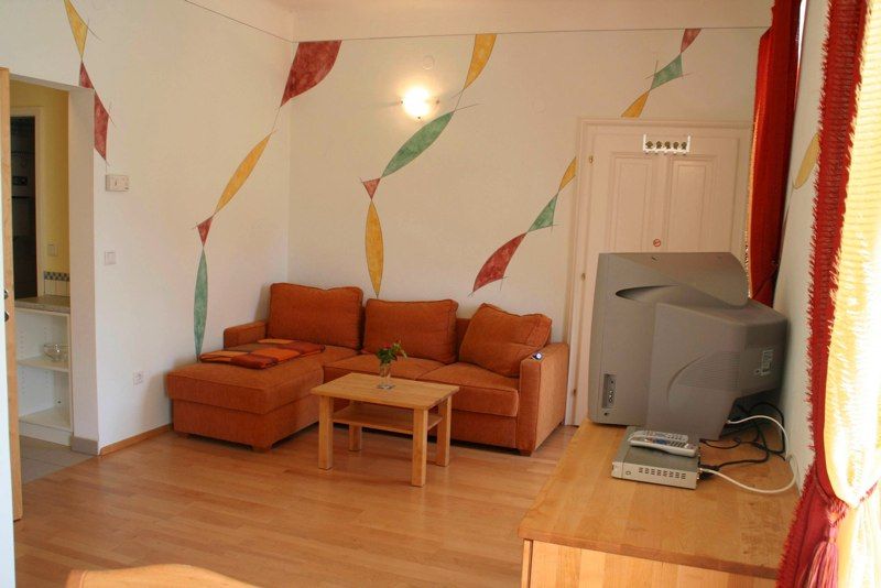 Obývacia izba s oranžovou pohovkou, dreveným stolom, TV a farebnými nástennými dekoráciami.