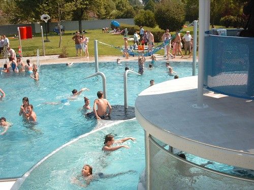 Ľudia sa môžu kúpať a hrať vo vonkajšom bazéne so šmykľavkou a zelenou plochou.