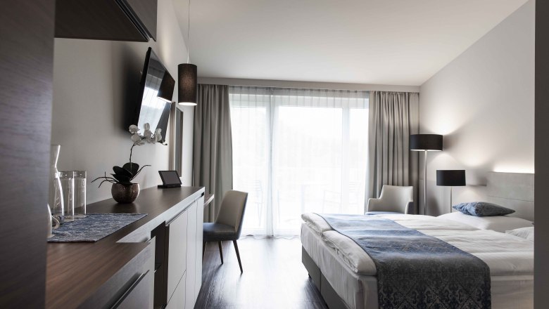 Modern&aacute; hotelov&aacute; izba s manželskou posteľou, TV, p&iacute;sac&iacute;m stolom a veľk&yacute;mi oknami so z&aacute;vesmi.