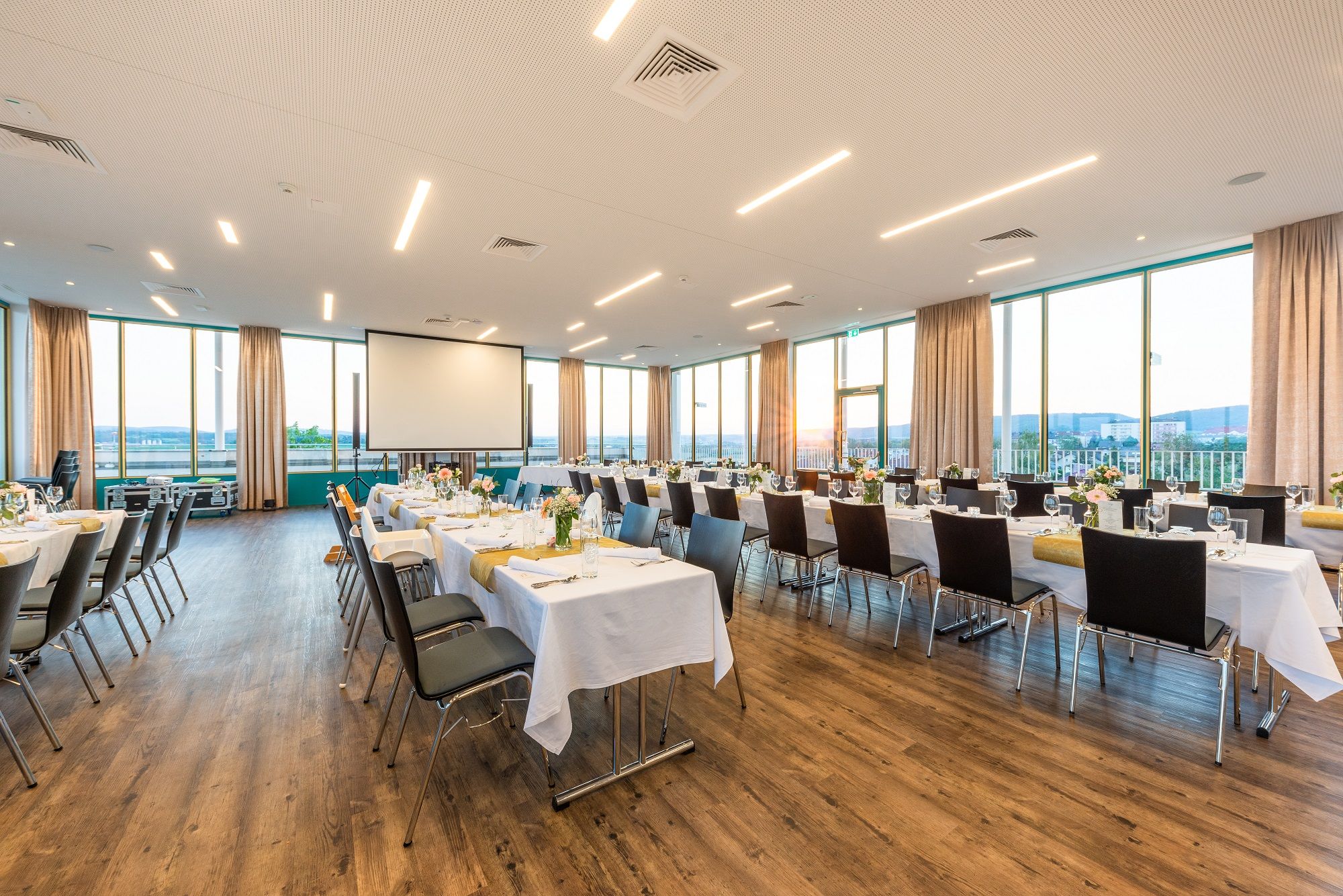 Veľká, svetlá miestnosť s krytými stolmi a stoličkami, veľkými oknami a projektorom.