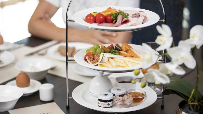 Raňajkov&yacute; st&ocirc;l s et&aacute;žou plnou syrov, &uacute;den&iacute;n, ovocia a n&aacute;tierok. V popred&iacute; biela orchidea.