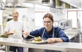 Šéfkuchár v reštauračnej kuchyni pripravuje jedlo, zatiaľ čo jeho kolega v pozadí drží iné jedlo.