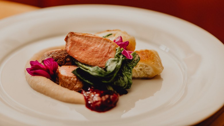 Ružov&aacute; pečen&aacute; kačica, kr&eacute;mov&eacute; kol&aacute;čiky, ga&scaron;tanov&eacute; pyr&eacute; a zelen&eacute; listy, zdoben&eacute; jedl&yacute;mi kvetmi.