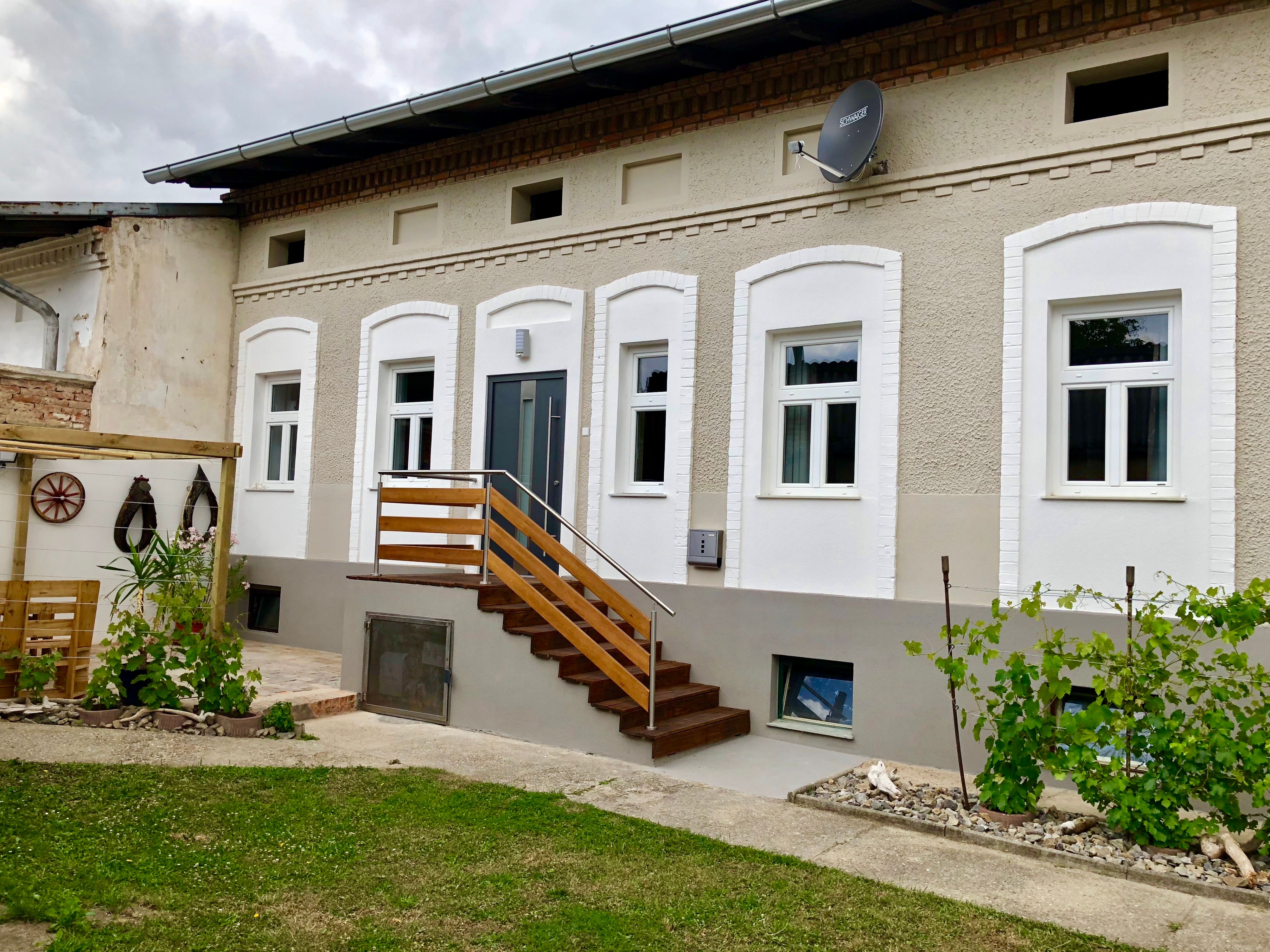 Zrekonštruovaný dom s bielymi oknami a dreveným schodiskom, obklopený malou záhradou s rastlinami.