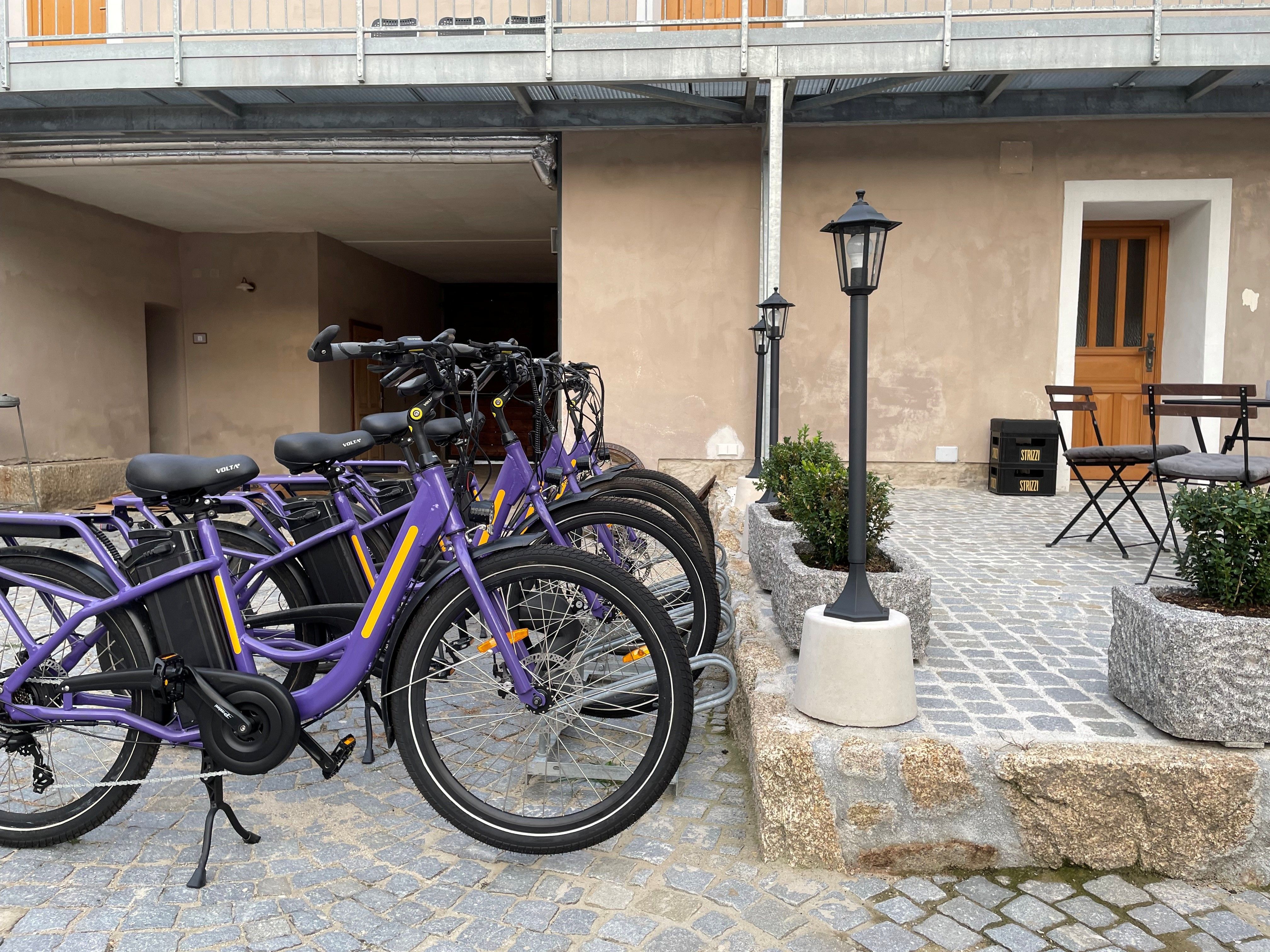 Na dláždenom dvore vedľa budovy s drevenými dverami a záhradným nábytkom stojí niekoľko fialových bicyklov.
