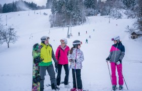 Skupina lyžiarov a snowboardistov na zasneženej zjazdovke s lesom v pozad&iacute;.