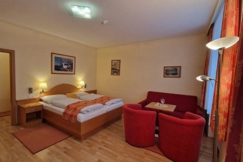 Hotelová izba s manželskou posteľou, červenou pohovkou a kreslami, drevenou podlahou a nástennými maľbami.