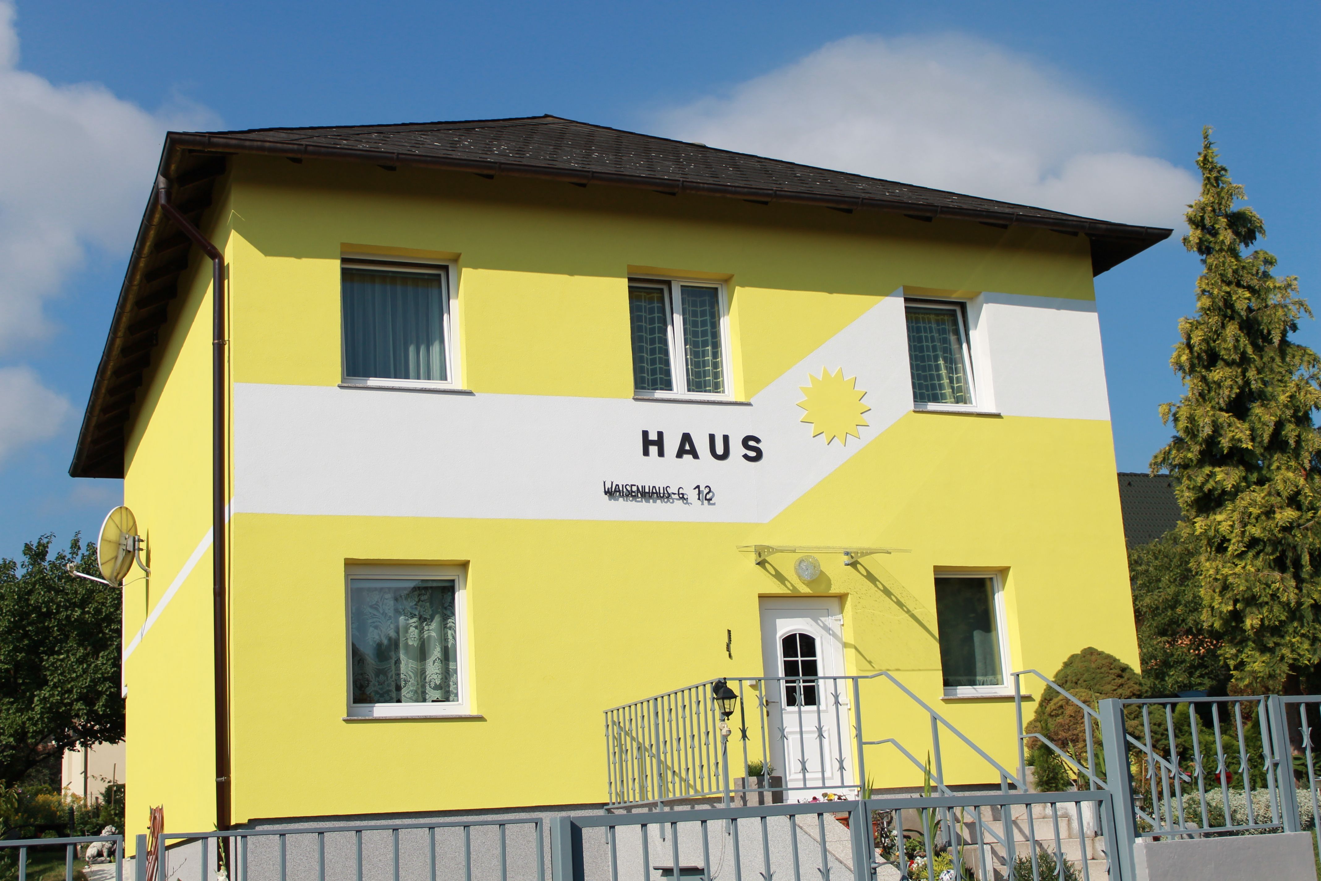 Žltý dom s nápisom "Haus" a slnkom na fasáde.