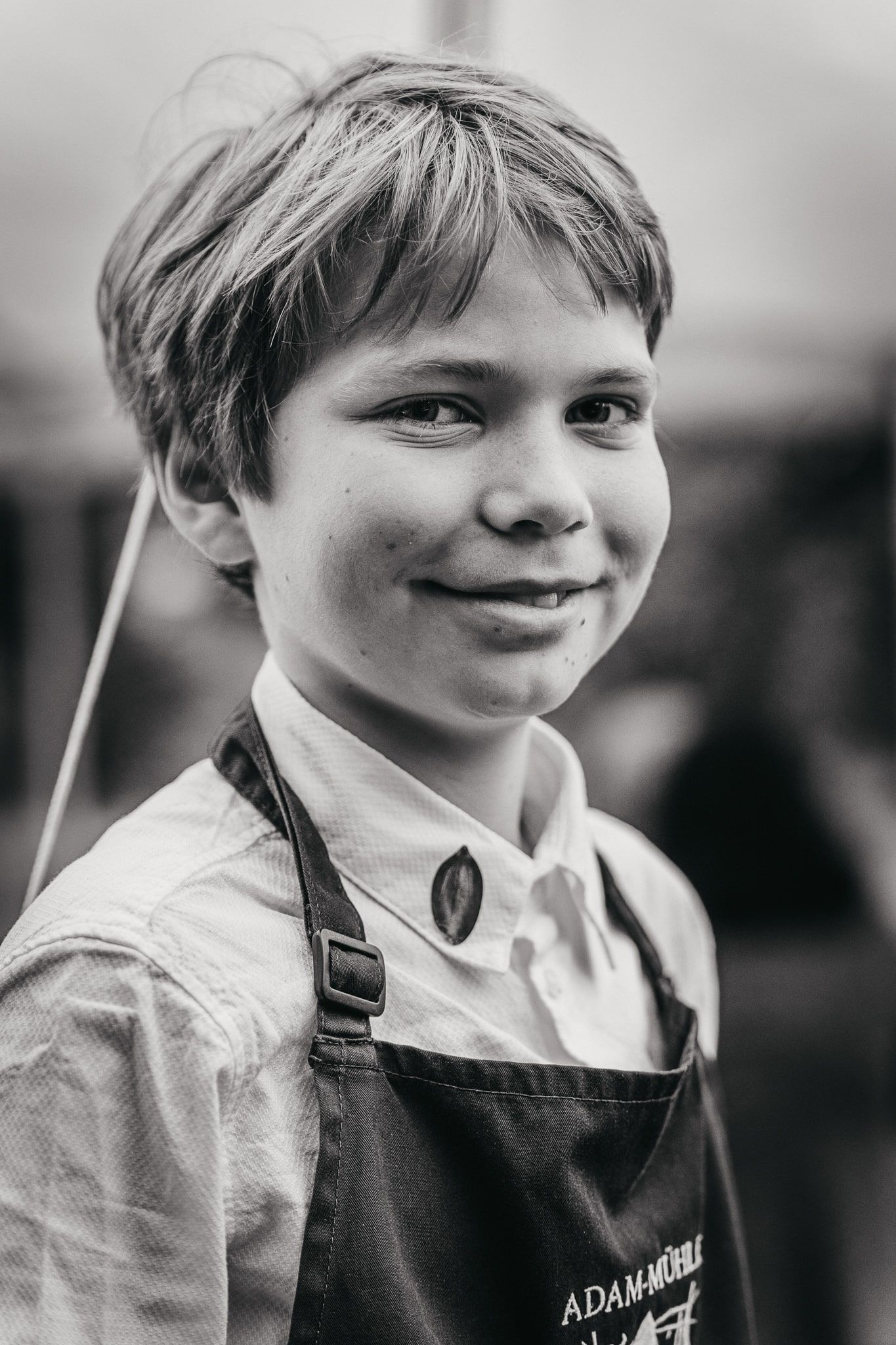 Chlapec v zástere s nápisom "Adam-Mühle" sa usmieva do kamery.