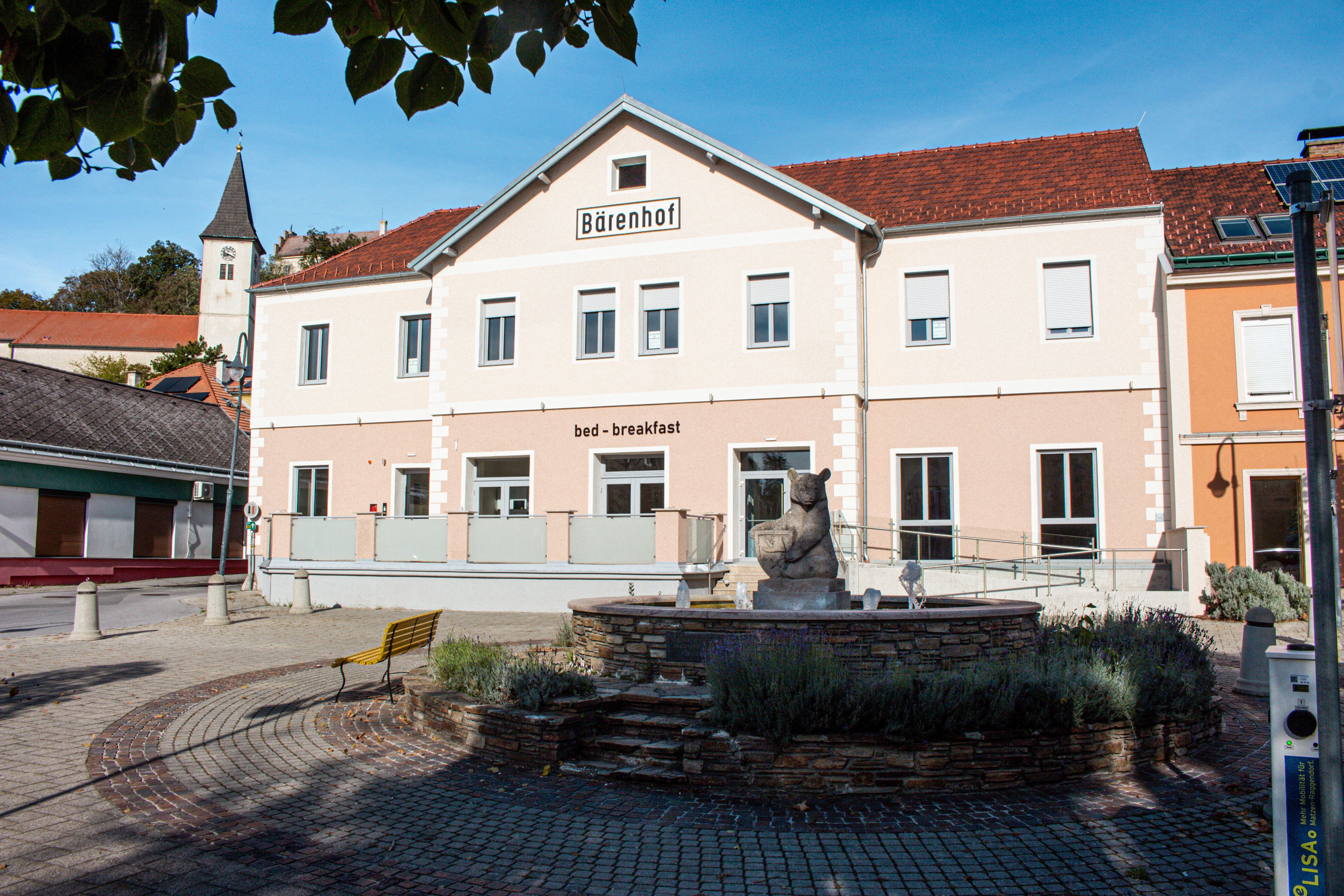 Budova s označením "Bärenhof" a "bed - breakfast" v slnečnom prostredí s fontánou v popredí.