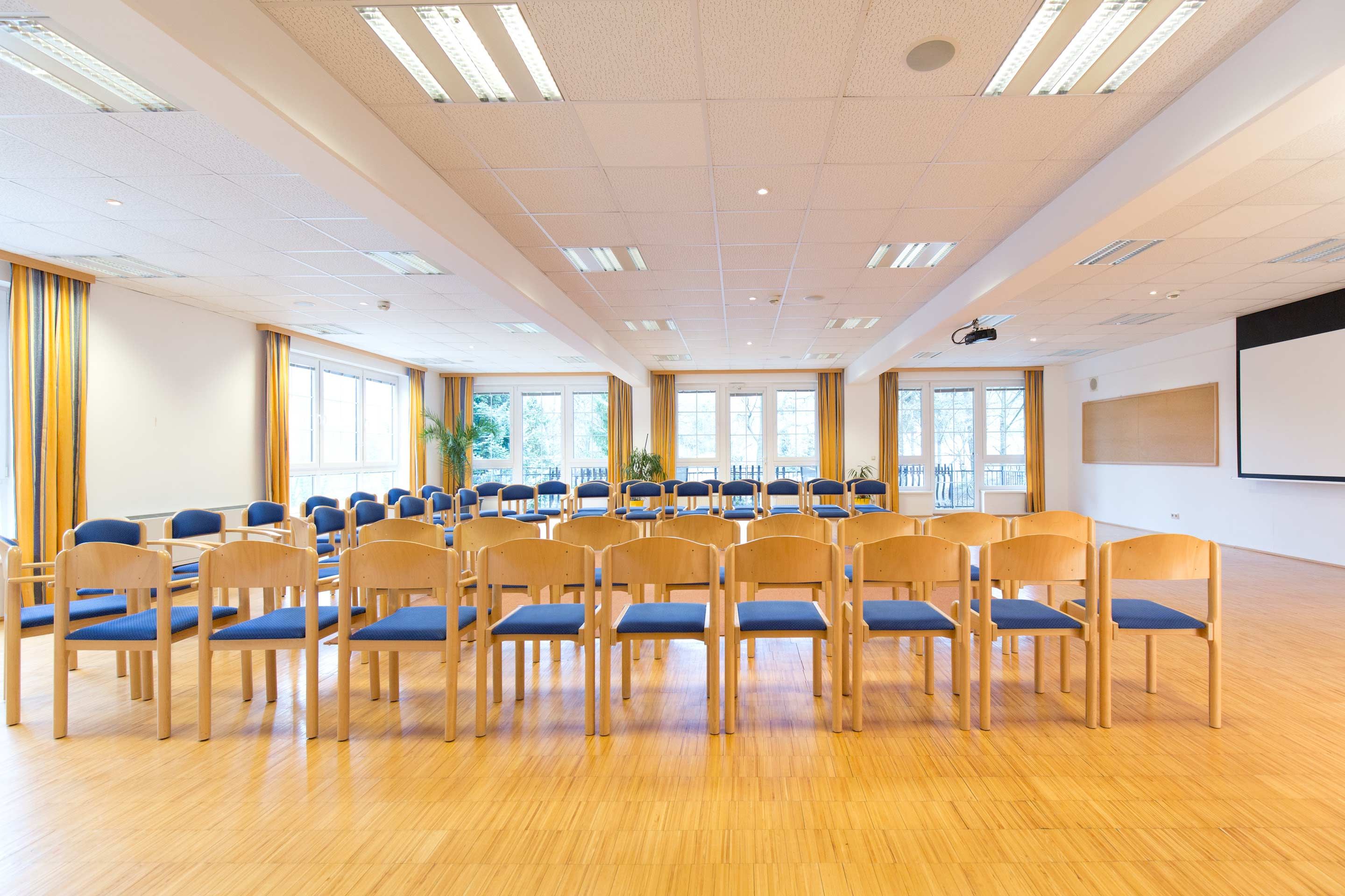 Svetlá seminárna miestnosť s radmi stoličiek, veľkými oknami a projektorom na strope.