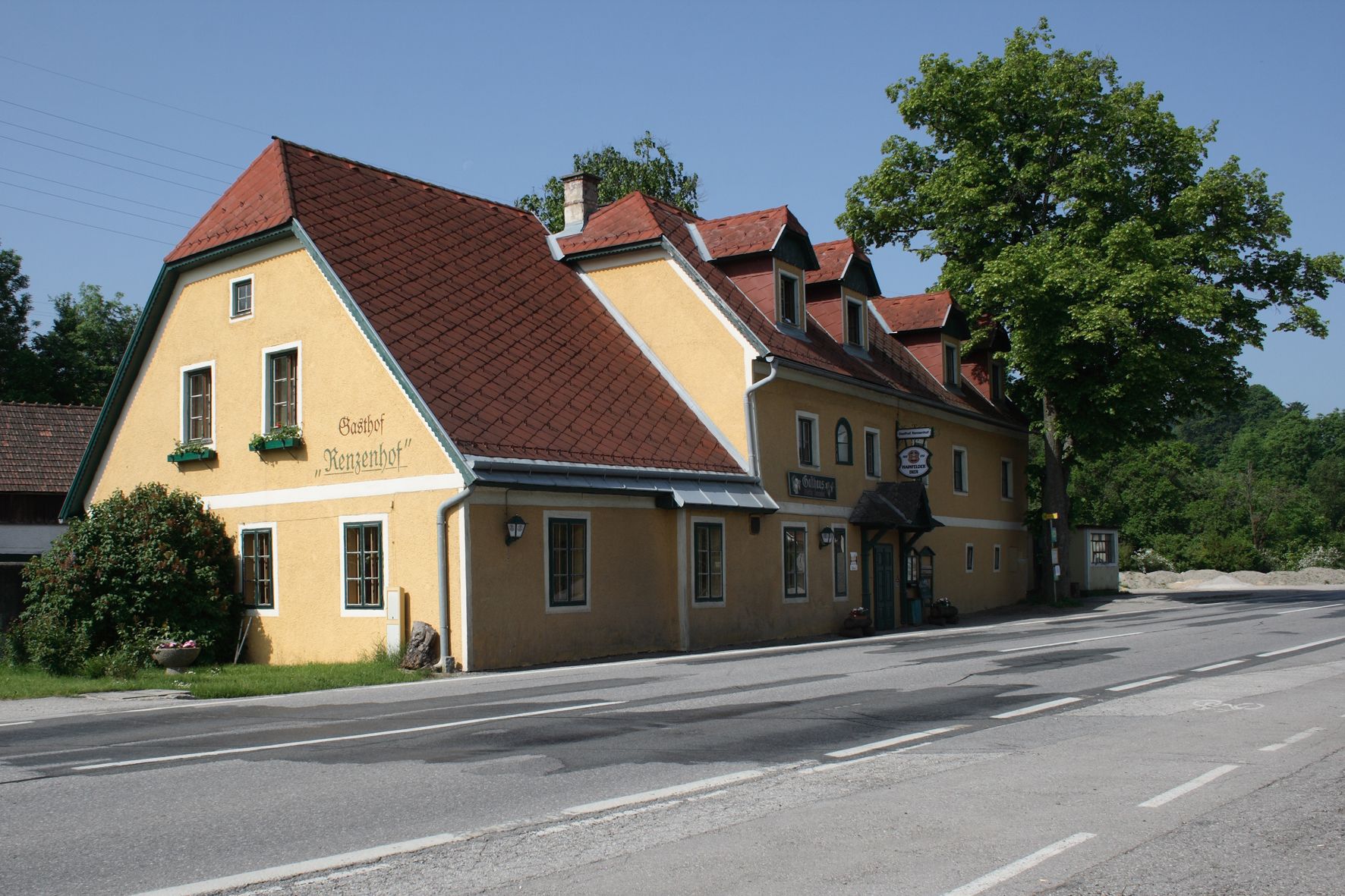 Žltý hostinec s červenou strechou na ceste, obklopený stromami.