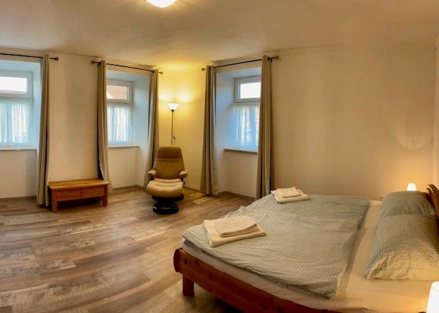 Jednoduchá spálňa s manželskou posteľou, kreslom, nočným stolíkom a tromi oknami so závesmi.