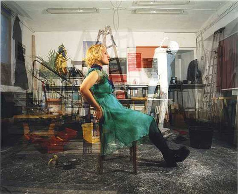 Žena v zelených šatách sedí na stoličke v preplnenej miestnosti.