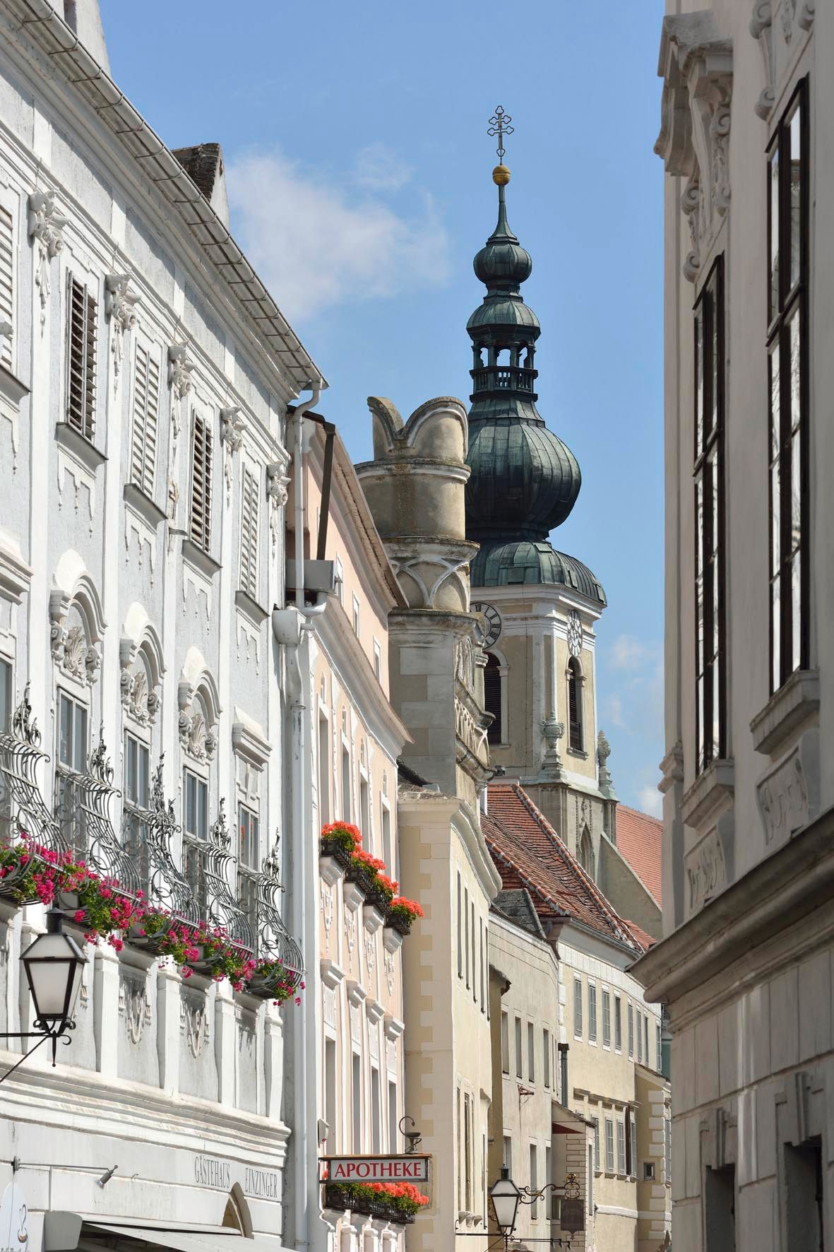 Scéna ulice s historickými budovami a kostolnou vežou v pozadí.