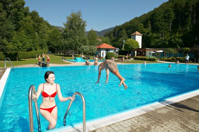 Vonkajší bazén, v ktorom sa ľudia kúpu a relaxujú. Žena vychádza z vody, zatiaľ čo muž skáče do bazéna. Obklopený stromami a kopcami.