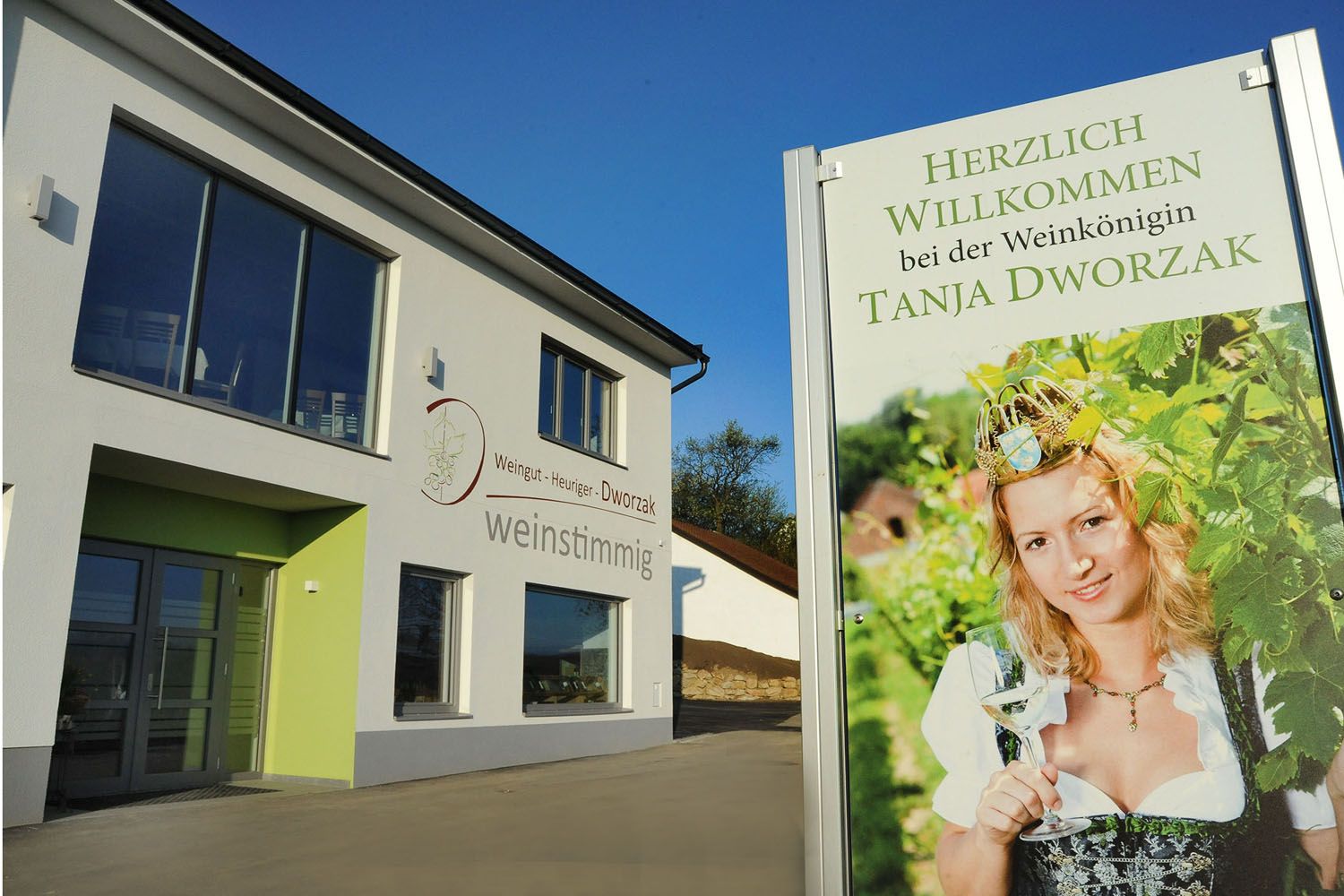 Moderná budova s nápisom zobrazujúcim ženu v tradičnom kroji s pohárom vína. Text: Vitajte u kráľovnej vína Tanji Dworzak.
