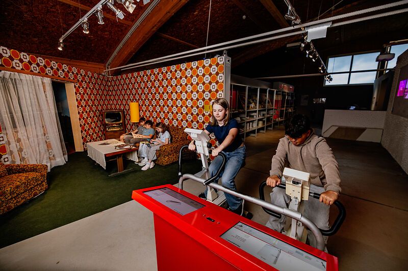 Mladí ľudia na bicyklových ergometroch v miestnosti s retro zariadením obývačky.