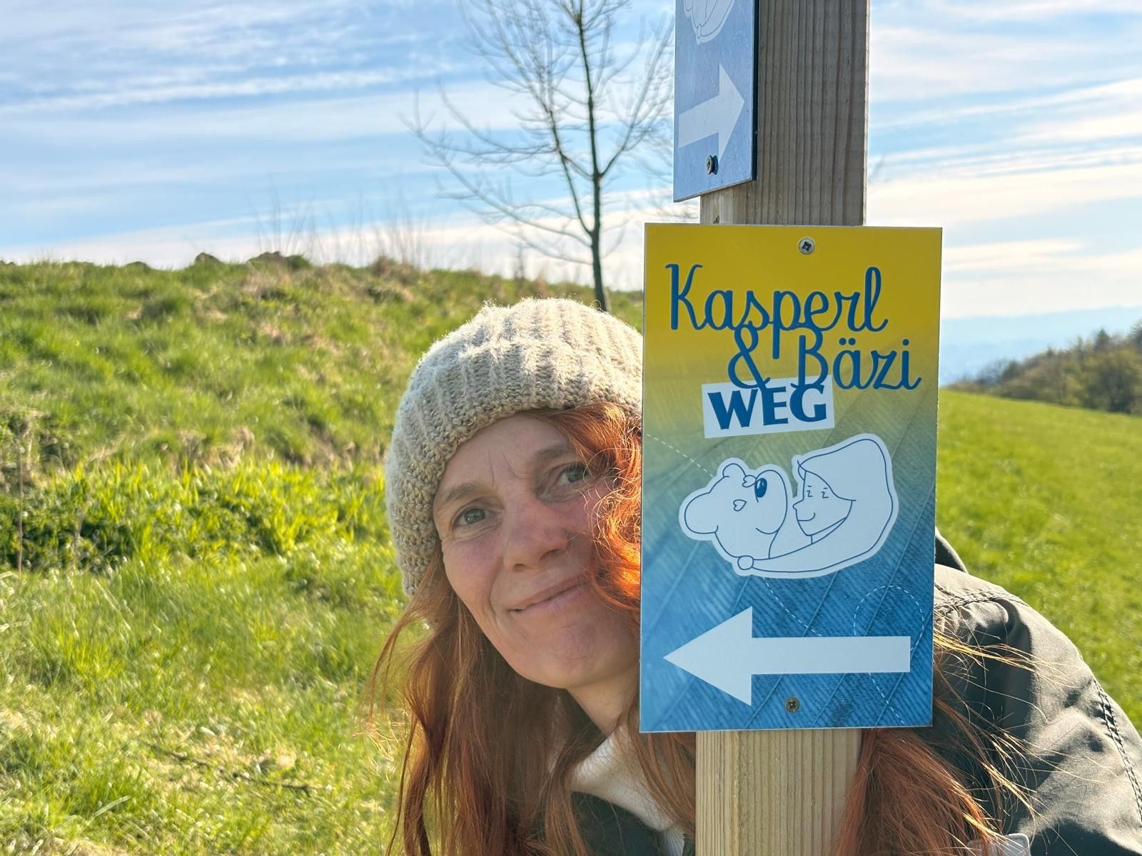 Žena v čiapke stojí vedľa smerovníka s nápisom „Kasperl & Bäzi Weg“ uprostred zelenej krajiny.