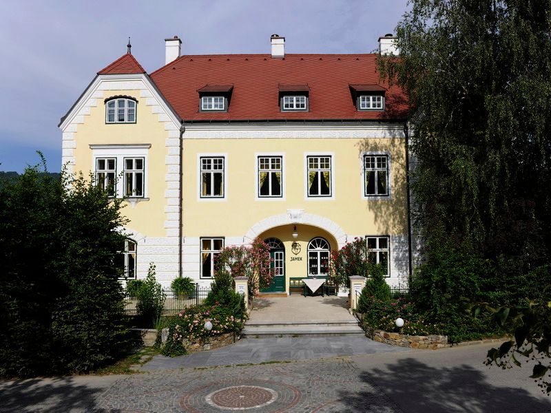 Žltá budova s červenou strechou a zelenými dverami, obklopená stromami a rastlinami.