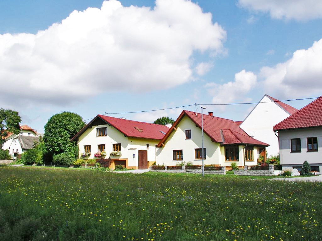 Ekologická farma s niekoľkými žltými budovami s červenými strechami, obklopená zelenými lúkami a stromami pod modrou oblohou s mrakmi.