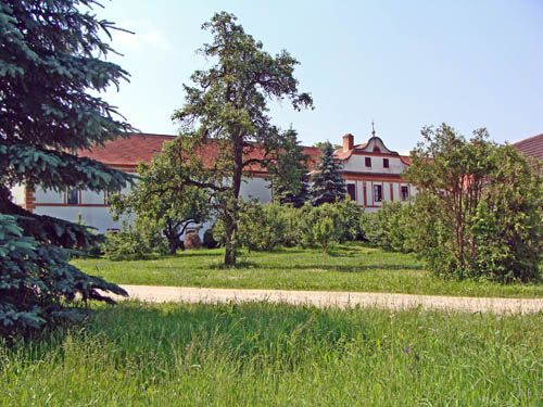 Historická budova s červenou strechou, obklopená stromami a zelenou lúkou.