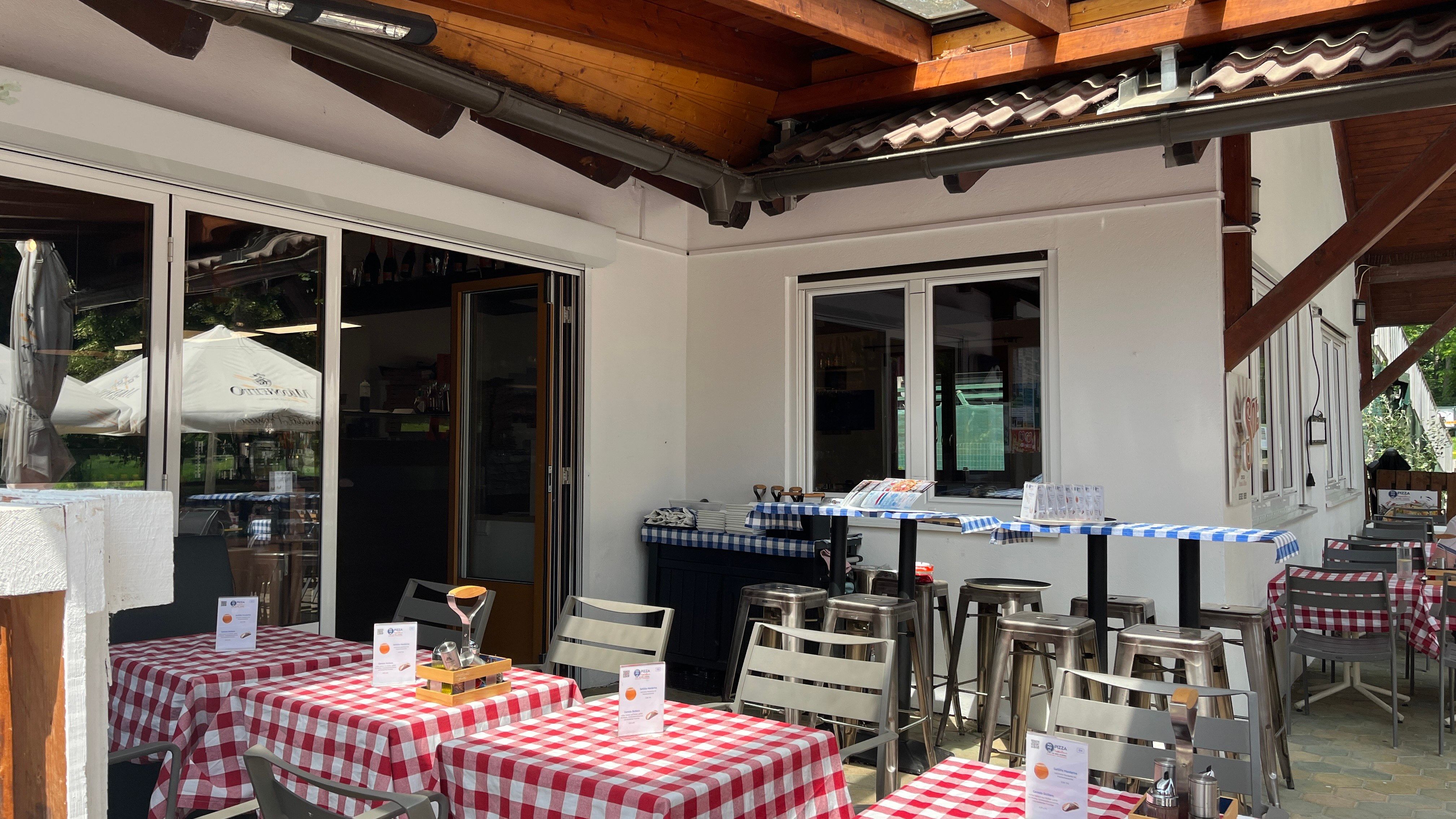 Vstupný priestor pizzerie so záhradou pre hostí. V popredí sú položené stoly s červenými obrusmi.