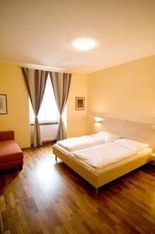 Útulná dvojlôžková izba s veľkou posteľou, drevenou podlahou, kreslom a oknom so závesmi.