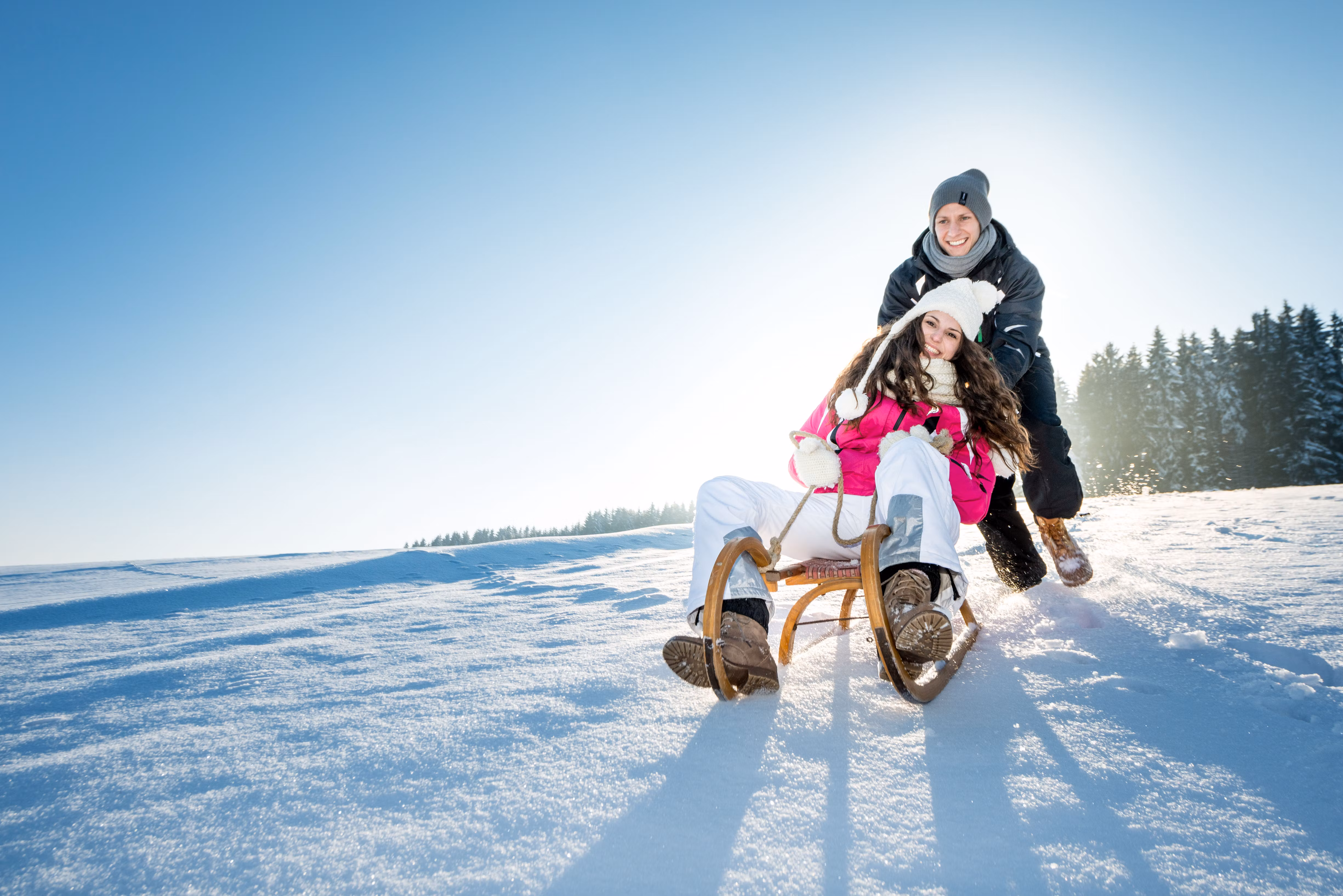 Ein fröhliches Paar genießt den Winter in der verschneiten Landschaft, während sie mit einem Schlitten den Hang hinunter sausen. Die strahlende Sonne und der glitzernde Schnee schaffen eine magische Atmosphäre, die zum Lachen und Spielen einlädt.