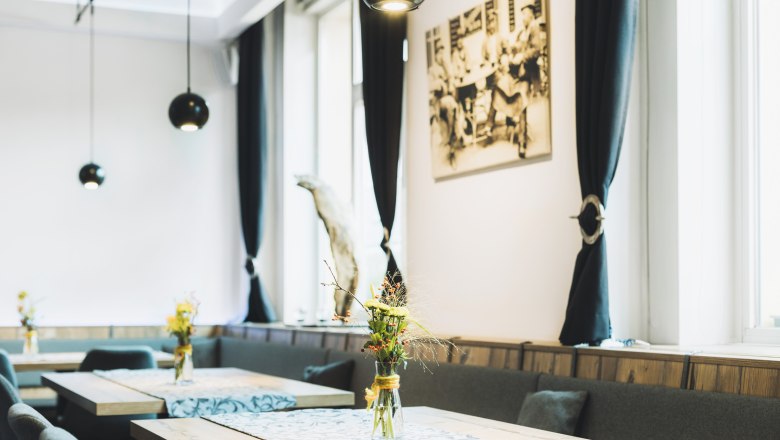 Pohľad do interiéru štýlovej kaviarne s drevenými stolmi, vázami na kvety a čiernymi závesmi.