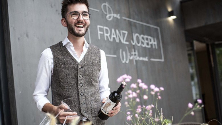Muž s okuliarmi a vo veste drží fľašu vína pred reštauráciou "Franz Joseph".