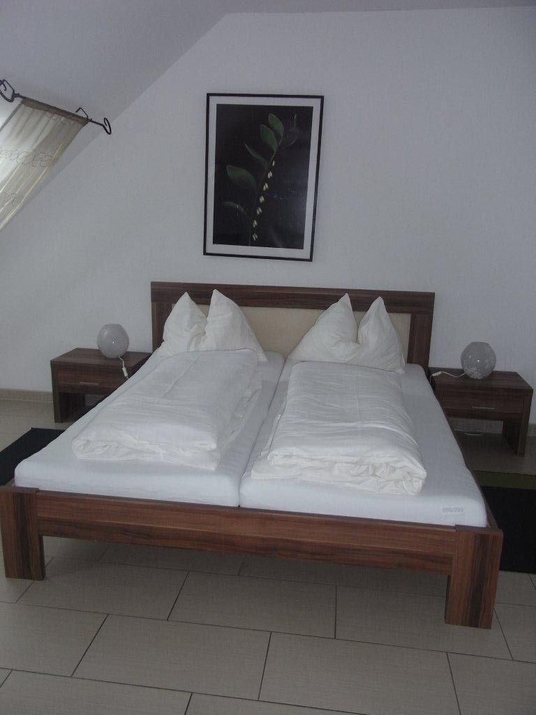 Manželská posteľ v hotelovej izbe s bielou posteľnou bielizňou a obrázkom rastliny na stene.