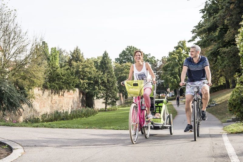 Dvojica jazdí na bicykloch po ceste v parku. Žena ťahá detský príves.