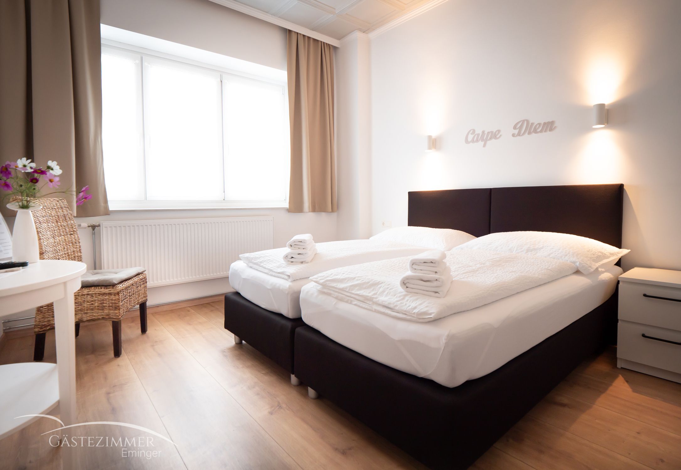 Moderná dvojlôžková izba s dvoma posteľami, bielymi perinami a uterákmi. Nápis "Carpe Diem" na stene, svetlé závesy a drevená podlaha.