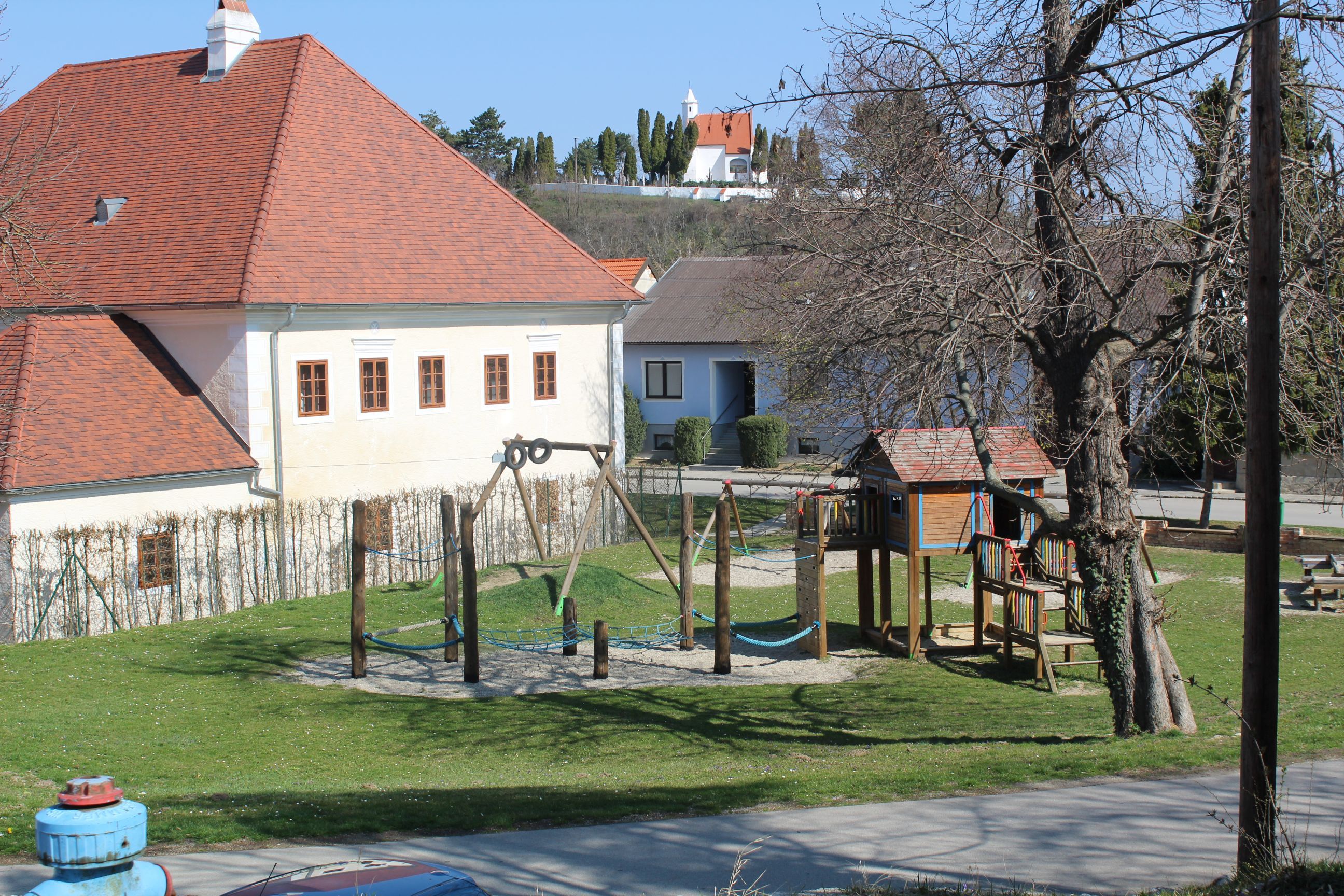 Detské ihrisko s preliezačkami a hojdačkami vedľa budovy s červenou strechou.