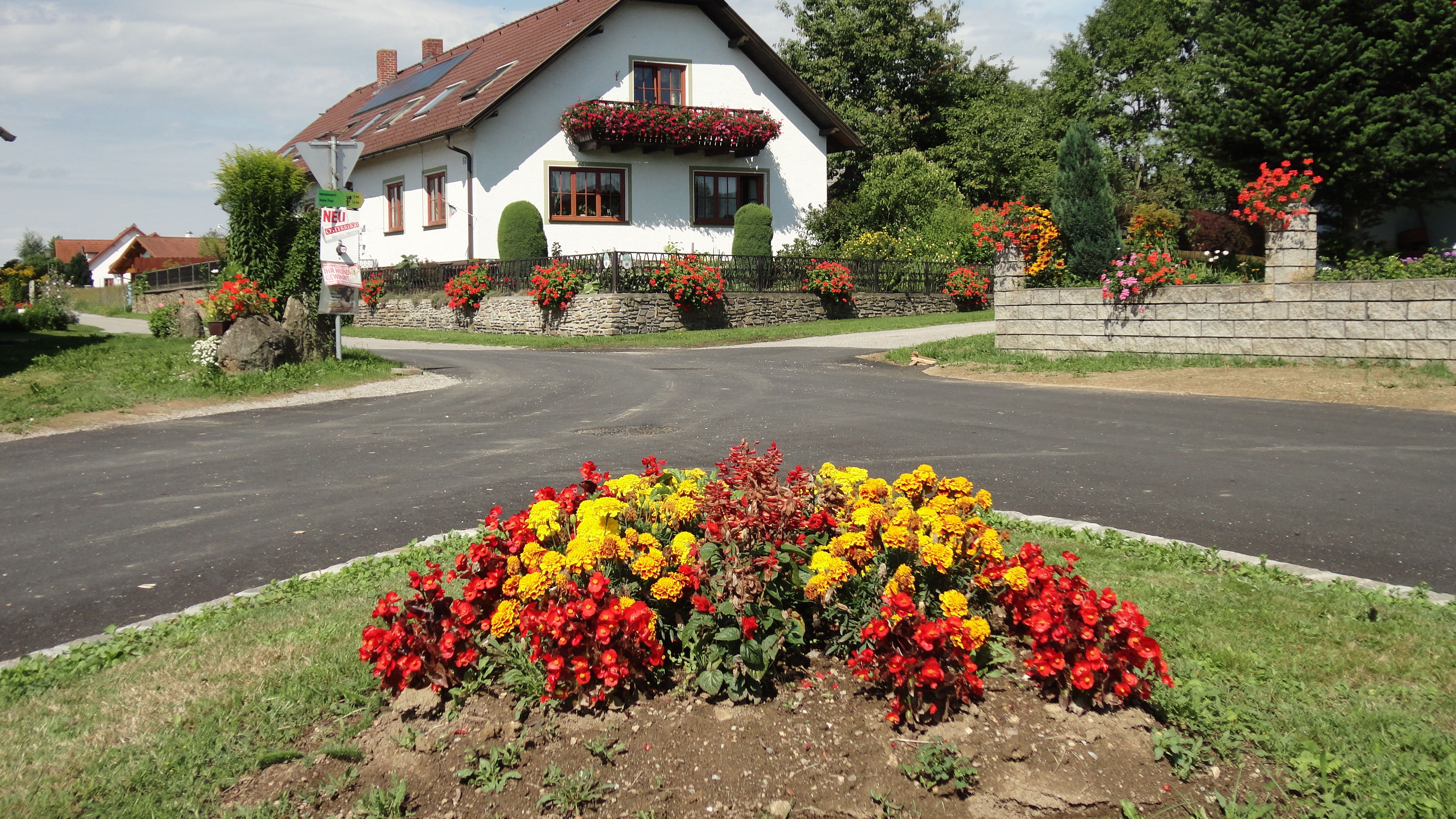 Biely dom s červenými škridlami a kvetinovou výzdobou, obklopený farebnými záhonmi a zelenou krajinou.